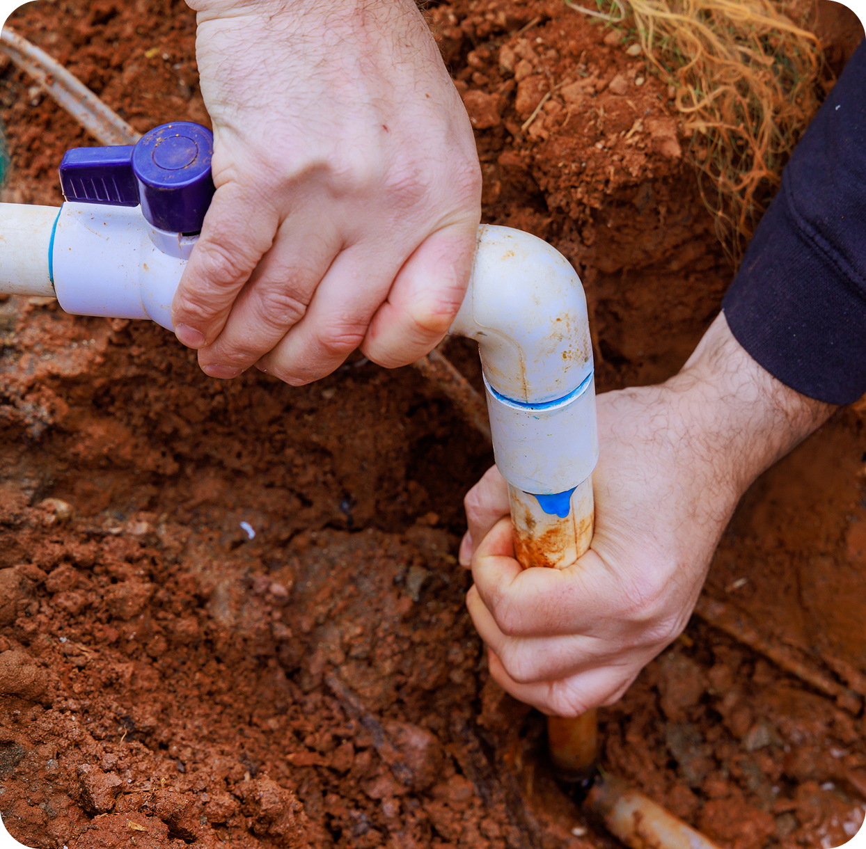 In a demonstration of community water services, a person carefully fixes a white PVC pipe with a blue valve connector embedded in the rich red soil.