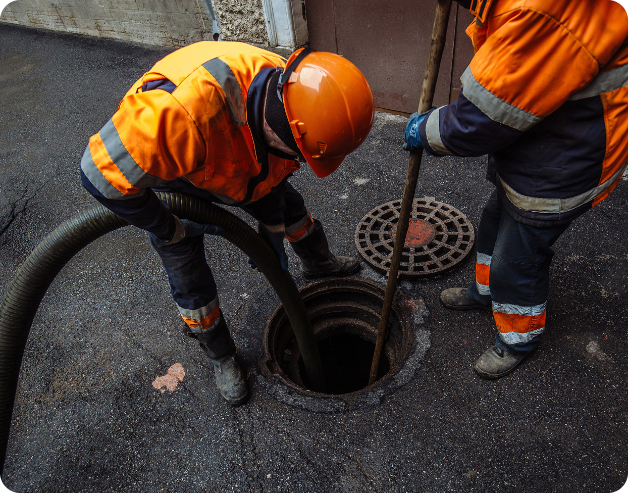 Two workers in orange safety gear use equipment to clean a manhole on a paved street, ensuring the smooth operation of Community Water Services.